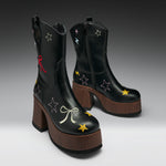 A pair of black leather ankle boots with brown platform heels, decorated with embroidered stars, bows, and lightning bolts in multiple colors, shown against a dark gray gradient background.