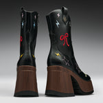 A pair of black leather mid-calf boots with chunky brown wooden platform heels, decorated with colorful embroidered stars, lightning bolts, and bows on a gray gradient studio background.