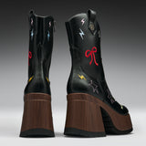 A pair of black leather mid-calf boots with chunky brown wooden platform heels, decorated with colorful embroidered stars, lightning bolts, and bows on a gray gradient studio background.