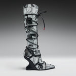 A single tall knee-high boot with a black and white manga comic book print, wrapped in black lace up to the ankle, set against a dark-to-light gray gradient studio background.