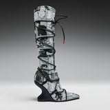 A single tall knee-high boot with a black and white manga comic book print, wrapped in black lace up to the ankle, set against a dark-to-light gray gradient studio background.