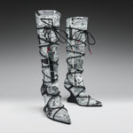A pair of thigh-high pointed-toe boots with a black and white newspaper collage print, wrapped in black lace up to the ankles, shown on a gray gradient studio background.