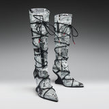 A pair of thigh-high pointed-toe boots with a black and white newspaper collage print, wrapped in black lace up to the ankles, shown on a gray gradient studio background.