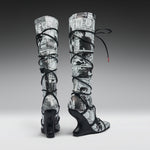 A pair of high-heeled, over-the-knee boots featuring a black and white newspaper print pattern wrapped around the shafts, with black lace-up straps and wedge heels, shown against a gray gradient studio background.