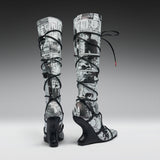 A pair of high-heeled, over-the-knee boots featuring a black and white newspaper print pattern wrapped around the shafts, with black lace-up straps and wedge heels, shown against a gray gradient studio background.