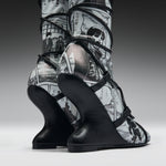 A pair of high-heeled boots with sculptural black wedge heels and black straps, featuring a black-and-white manga comic book print on the uppers, shown against a dark gray studio background.