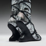 A pair of high-heeled boots with sculptural black wedge heels and black straps, featuring a black-and-white manga comic book print on the uppers, shown against a dark gray studio background.