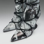 A pair of pointed-toe high-heeled boots featuring a black and white manga comic book print with black lace-up wrap details, shown on a neutral gray background.