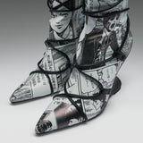 A pair of pointed-toe high-heeled boots featuring a black and white manga comic book print with black lace-up wrap details, shown on a neutral gray background.
