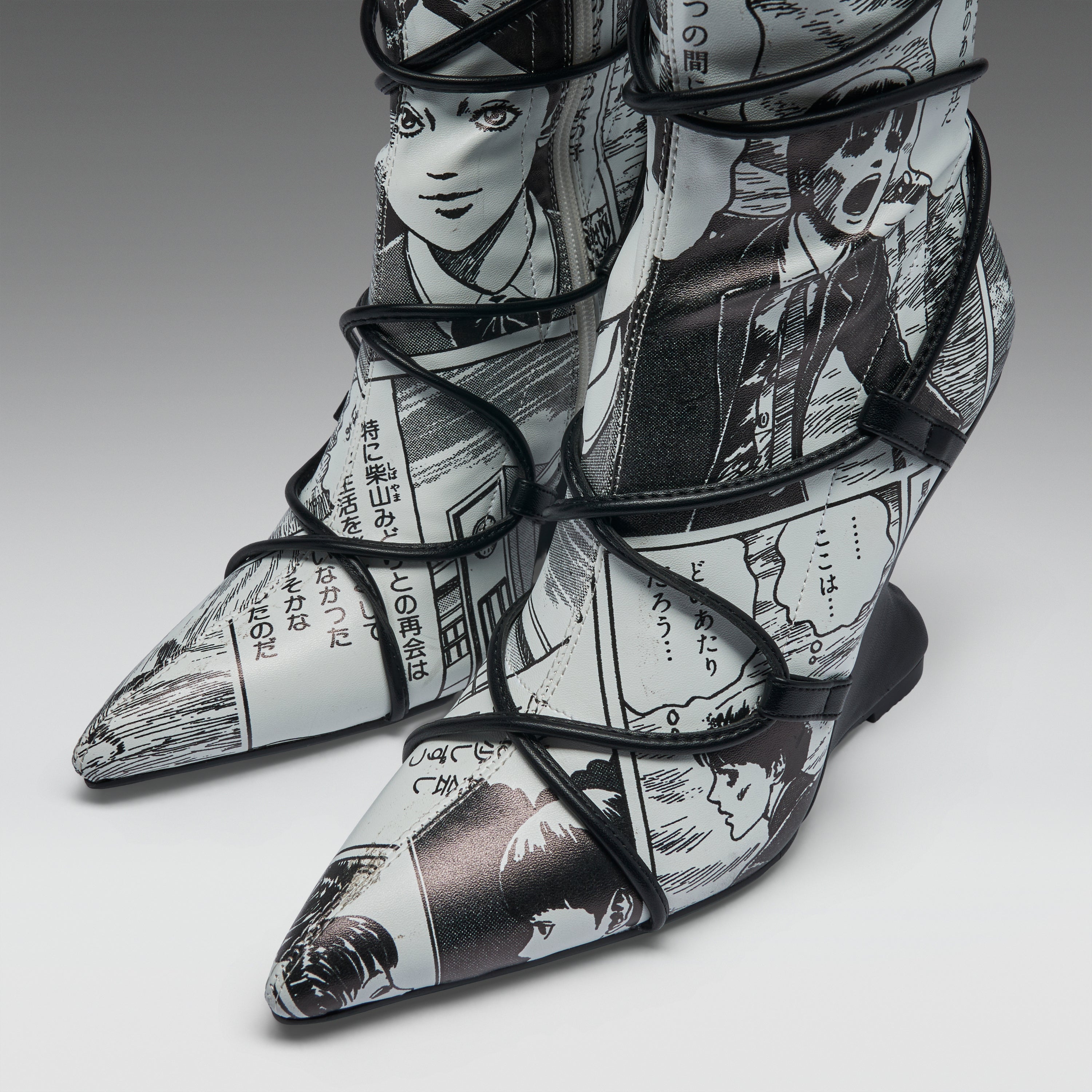 A pair of pointed-toe high-heeled boots featuring a black and white manga comic book print with black lace-up wrap details, shown on a neutral gray background.