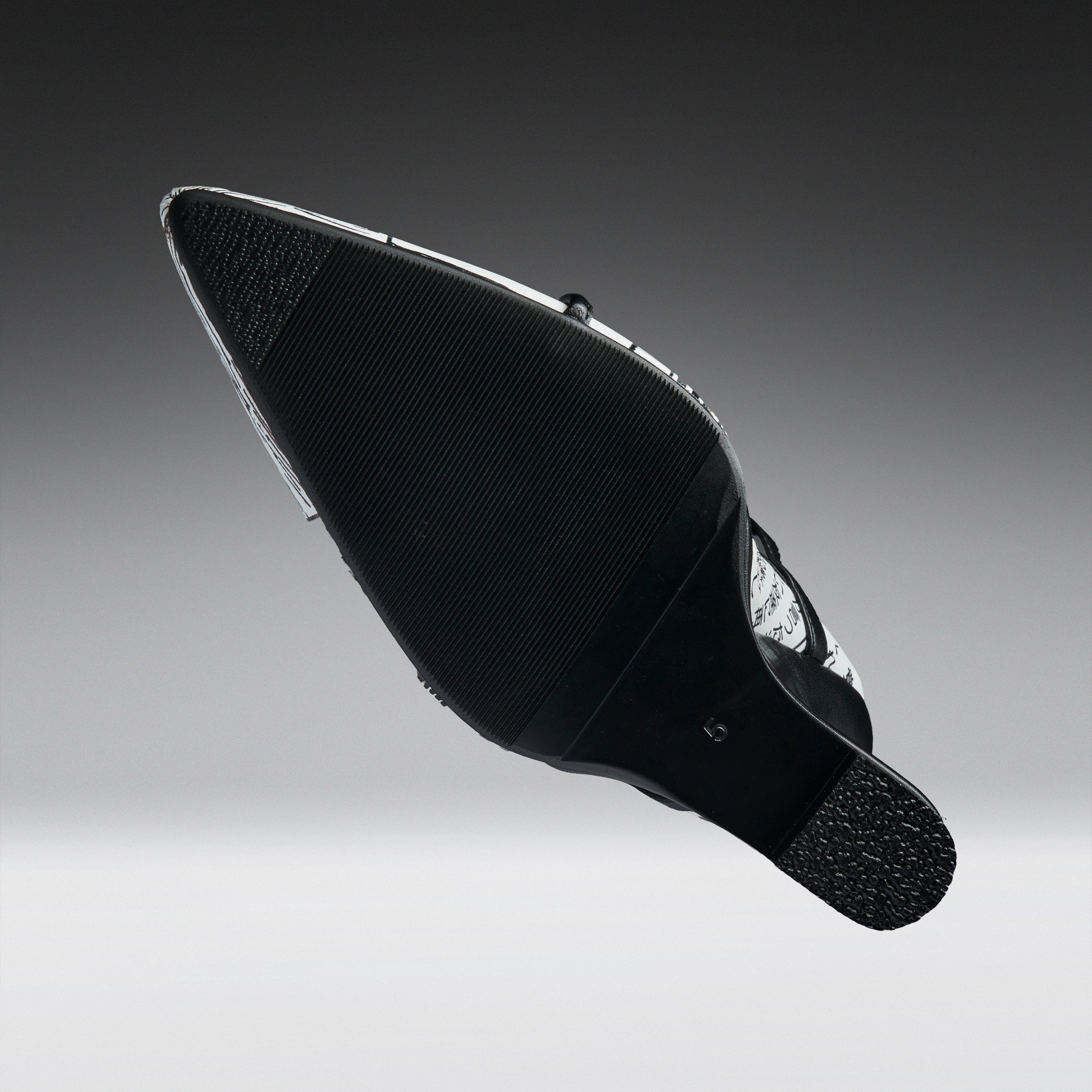 A black pointed-toe shoe shown from the sole side, featuring a textured black rubber outsole and a silver metallic side accent, set against a dark-to-light gray gradient background.