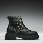 A black leather ankle boot with three silver buckle straps, white contrast stitching, and a chunky lug sole, shown against a dark gray gradient background.