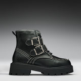 A black leather ankle boot with three silver buckle straps, white contrast stitching, and a chunky lug sole, shown against a dark gray gradient background.