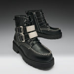 A pair of black leather lace-up ankle boots with silver buckle straps, white contrast stitching, and chunky black soles, shown on a dark-to-light gray gradient studio background.