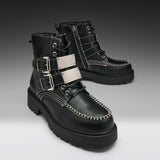 A pair of black leather lace-up ankle boots with silver buckle straps, white contrast stitching, and chunky black soles, shown on a dark-to-light gray gradient studio background.