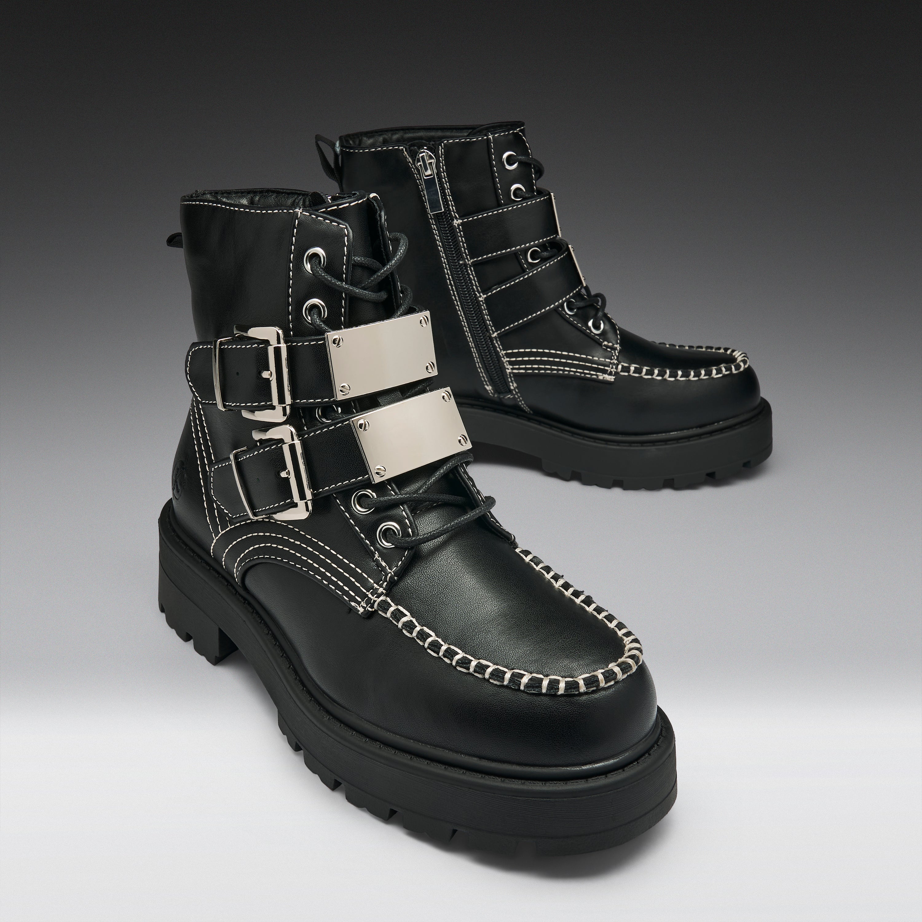 A pair of black leather lace-up ankle boots with silver buckle straps, white contrast stitching, and chunky black soles, shown on a dark-to-light gray gradient studio background.