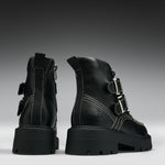A pair of black leather ankle boots with chunky lug soles, featuring silver buckle details, contrast white stitching, and side zippers, shown from the back and side against a dark gray gradient background.