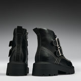 A pair of black leather ankle boots with chunky lug soles, featuring silver buckle details, contrast white stitching, and side zippers, shown from the back and side against a dark gray gradient background.