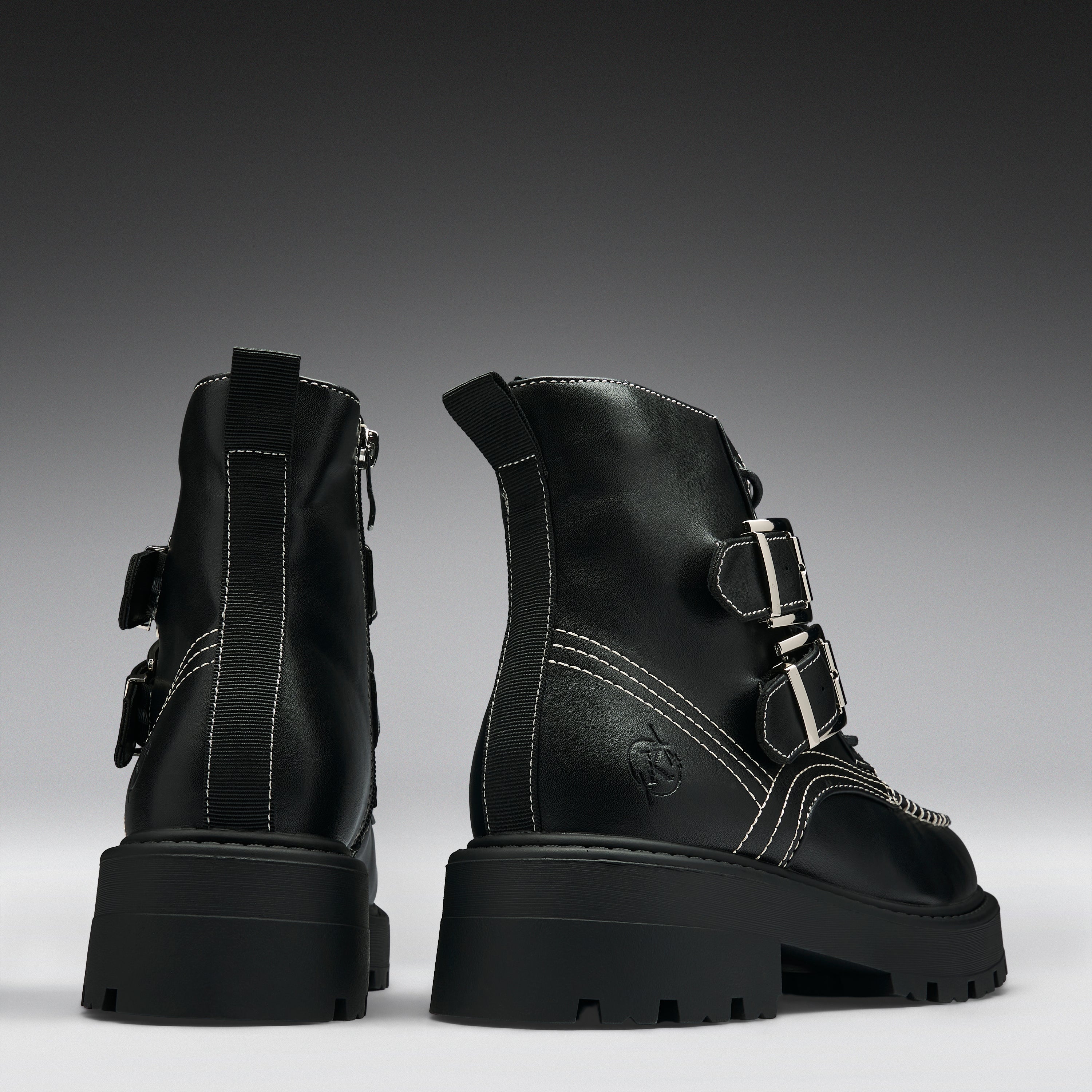 A pair of black leather ankle boots with chunky lug soles, featuring silver buckle details, contrast white stitching, and side zippers, shown from the back and side against a dark gray gradient background.