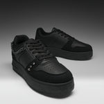 A pair of black low-top sneakers with matte and suede textures, featuring a chain detail on the side, metal studs, perforated detailing on the toe, and thick black soles, set against a dark gray gradient background.