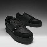 A pair of black low-top sneakers with matte and suede textures, featuring a chain detail on the side, metal studs, perforated detailing on the toe, and thick black soles, set against a dark gray gradient background.