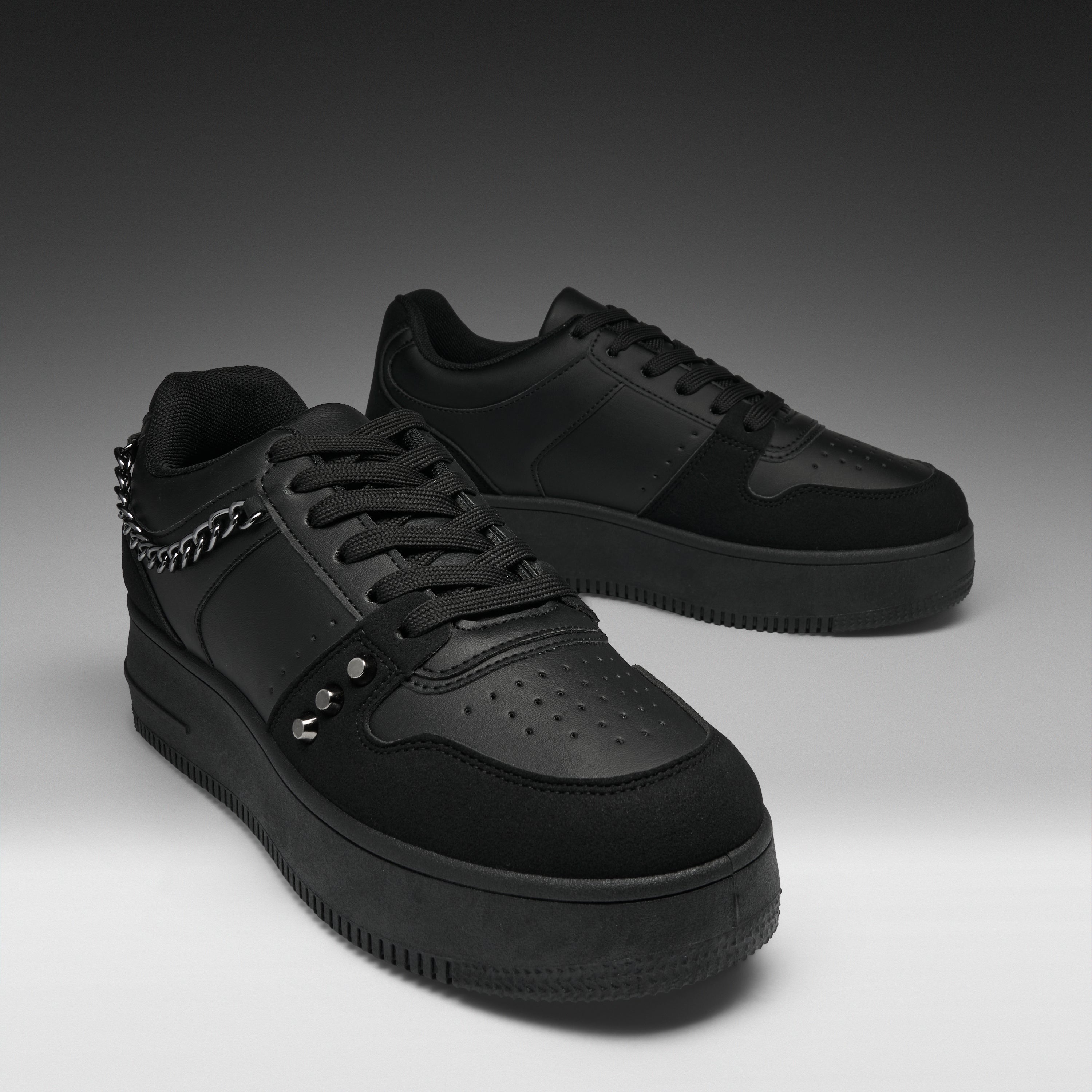 A pair of black low-top sneakers with matte and suede textures, featuring a chain detail on the side, metal studs, perforated detailing on the toe, and thick black soles, set against a dark gray gradient background.