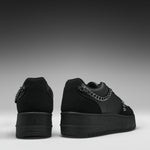 A pair of black low-top platform sneakers shown from the back, featuring a chunky black sole, black suede and leather panels, and a prominent silver chain detail across the back.