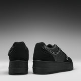 A pair of black low-top platform sneakers shown from the back, featuring a chunky black sole, black suede and leather panels, and a prominent silver chain detail across the back.