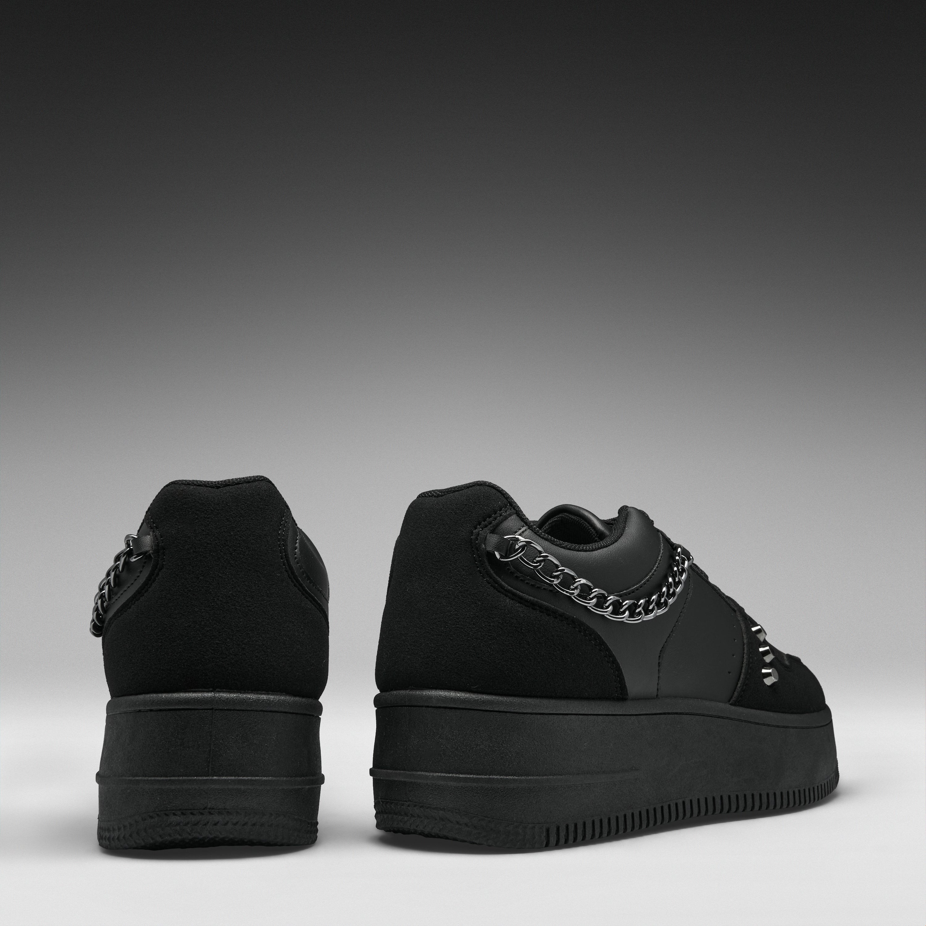 A pair of black low-top platform sneakers shown from the back, featuring a chunky black sole, black suede and leather panels, and a prominent silver chain detail across the back.