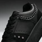A close-up of a black low-top sneaker with black laces, a thick black sole, and a prominent silver chain draped over the side near the collar.