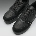 A pair of black low-top sneakers with perforated leather panels, black laces, and a small silver metal accent on the side, photographed on a clean gray studio background.
