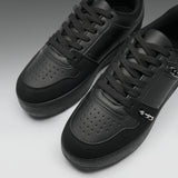 A pair of black low-top sneakers with perforated leather panels, black laces, and a small silver metal accent on the side, photographed on a clean gray studio background.