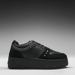 A black low-top platform sneaker with a chunky sole, featuring a silver chain detail across the side and three silver studs on the toe box, shown against a dark gray gradient background.
