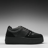 A black low-top platform sneaker with a chunky sole, featuring a silver chain detail across the side and three silver studs on the toe box, shown against a dark gray gradient background.