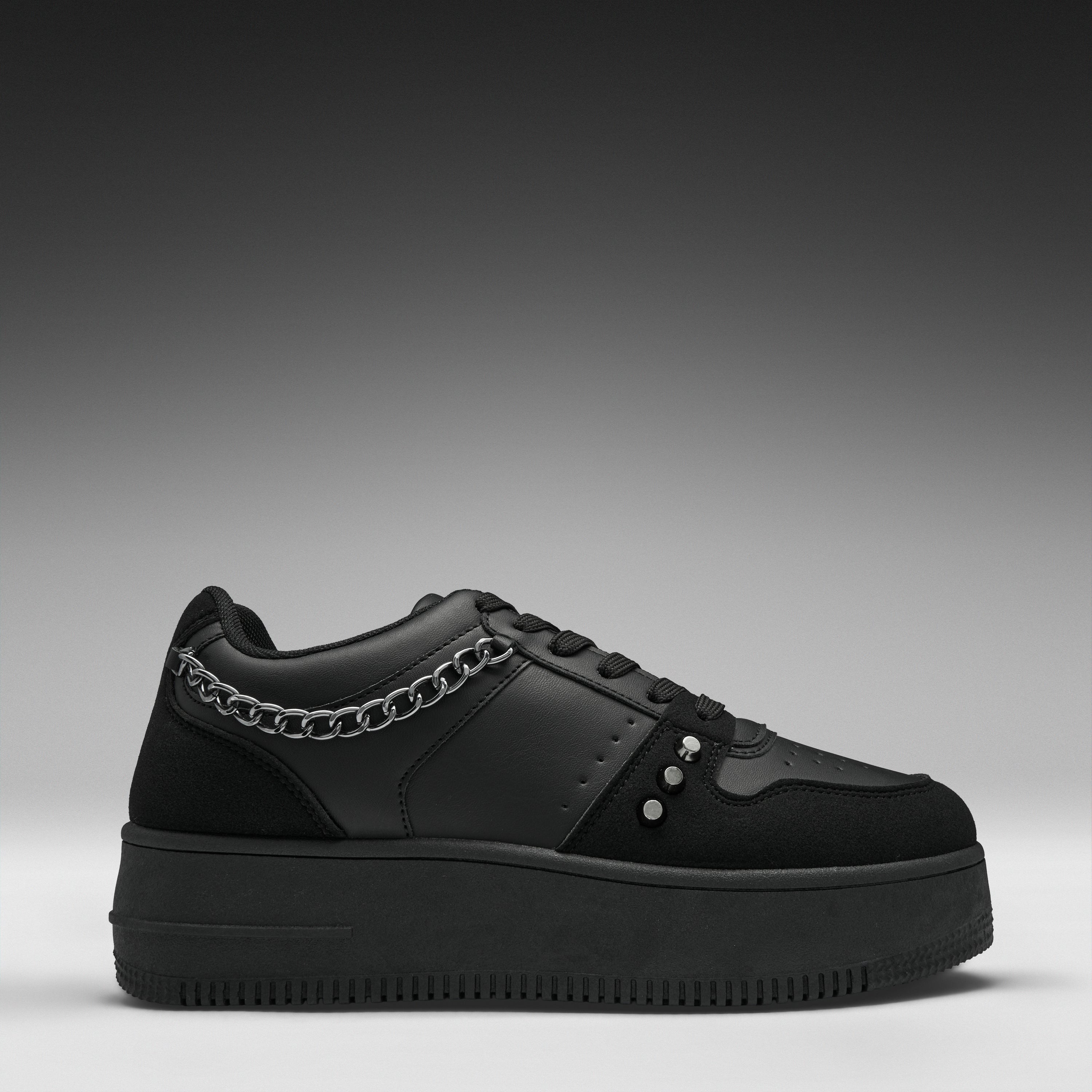 A black low-top platform sneaker with a chunky sole, featuring a silver chain detail across the side and three silver studs on the toe box, shown against a dark gray gradient background.