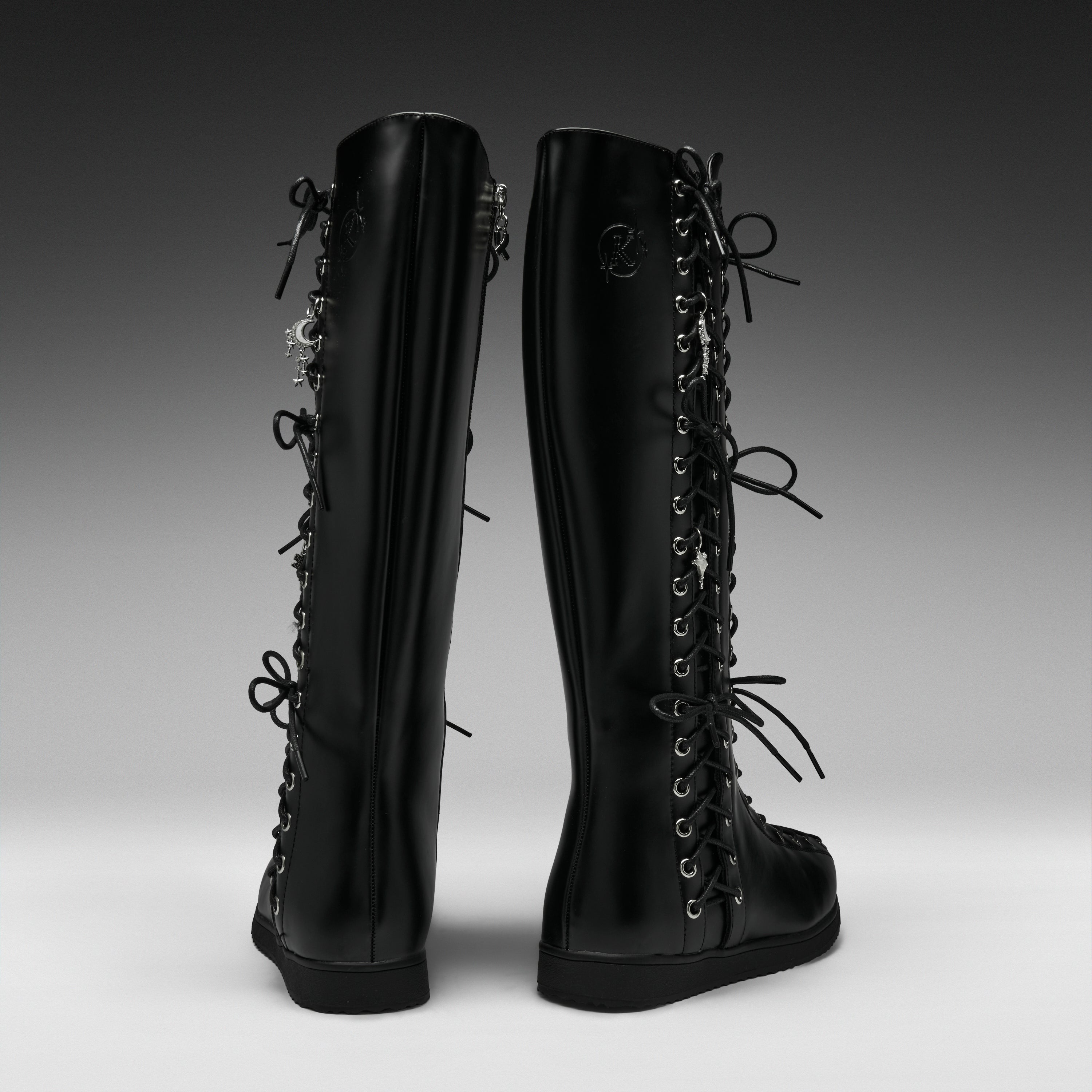 A pair of black knee-high lace-up boots shown from the back on a dark-to-light gray gradient studio background.