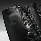 Close-up of a black leather lace-up boot with silver eyelets, a crescent moon and skull charm, and an embossed logo on the side.