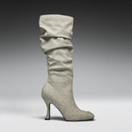A single knee-high slouch boot in light gray suede, featuring a pointed toe and a slim stiletto heel, shown against a dark-to-light gray studio gradient background.