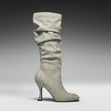 A single knee-high slouch boot in light gray suede, featuring a pointed toe and a slim stiletto heel, shown against a dark-to-light gray studio gradient background.