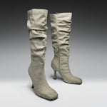 A pair of tall, knee-high heeled boots in a light taupe suede-like material, featuring a ruched shaft and a side zipper, set against a dark-to-light gray gradient studio background.