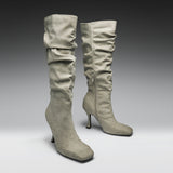 A pair of tall, knee-high heeled boots in a light taupe suede-like material, featuring a ruched shaft and a side zipper, set against a dark-to-light gray gradient studio background.