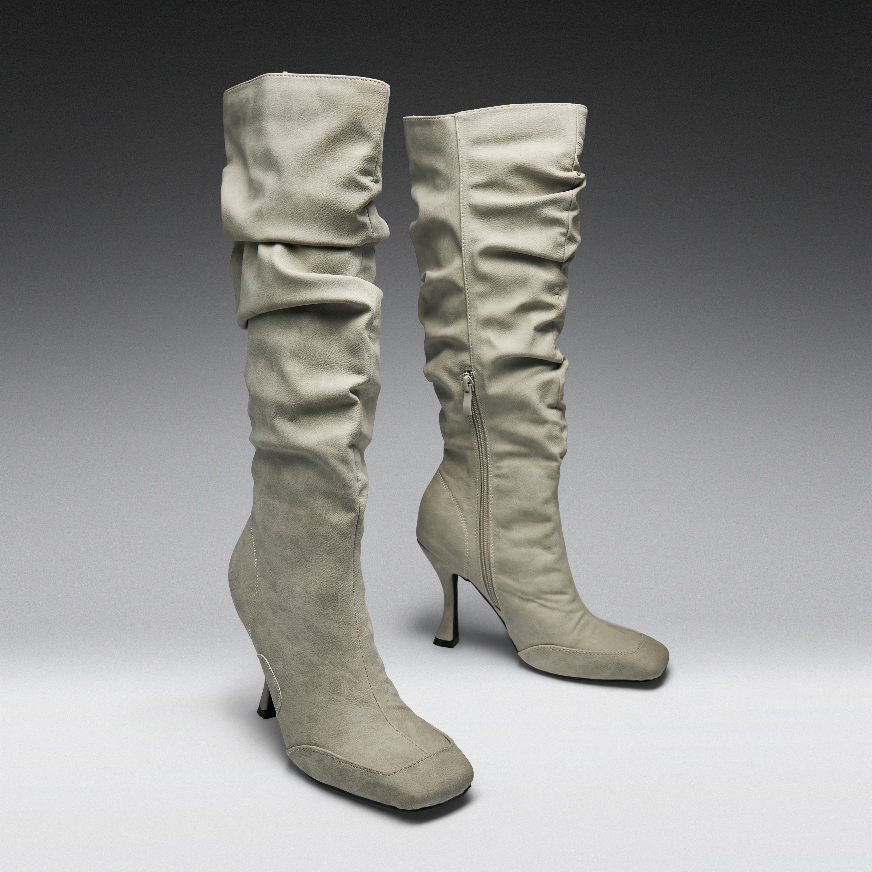 A pair of tall, knee-high heeled boots in a light taupe suede-like material, featuring a ruched shaft and a side zipper, set against a dark-to-light gray gradient studio background.