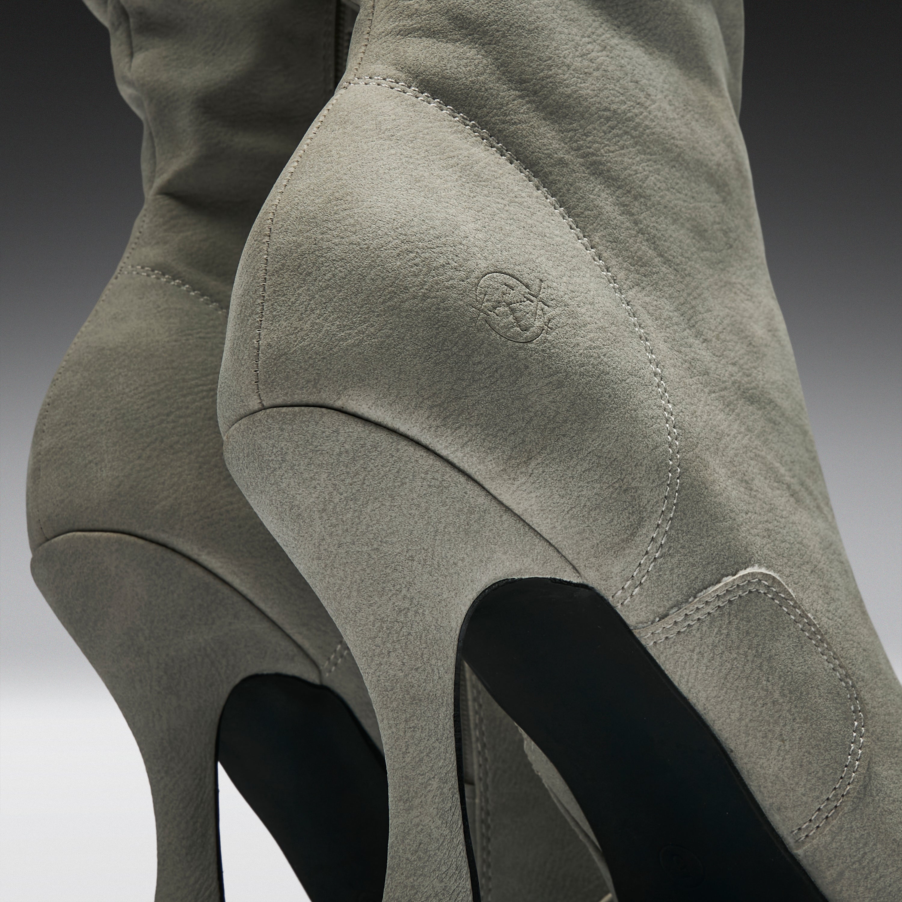 Close-up of a pair of over-the-knee boots in light gray suede, showing the side and back details, high stiletto heels, and a small debossed logo on the side.