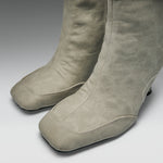 A pair of tall taupe suede boots with a square toe and low block heel, shown from a close-up angle on a neutral gray background.