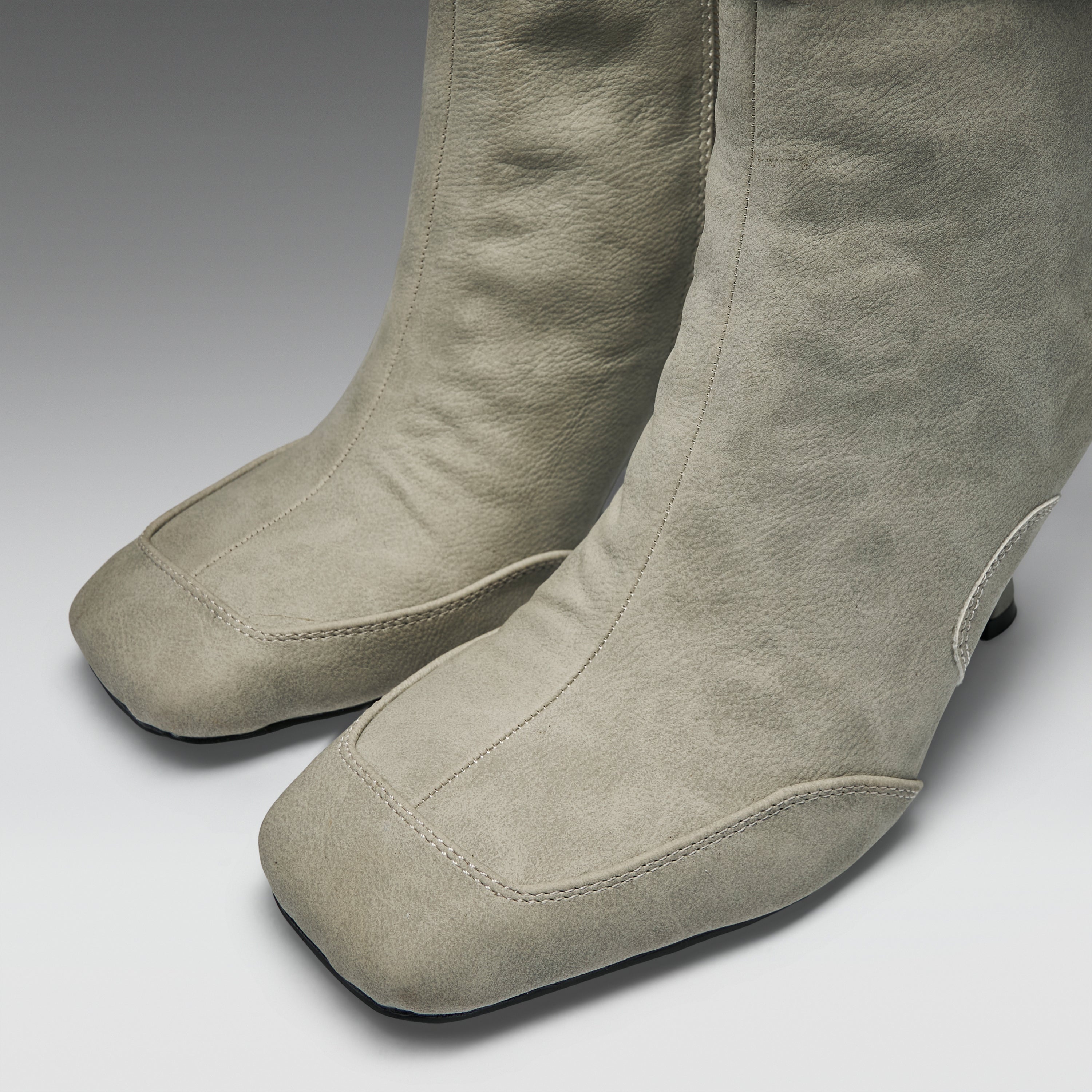 A pair of tall taupe suede boots with a square toe and low block heel, shown from a close-up angle on a neutral gray background.
