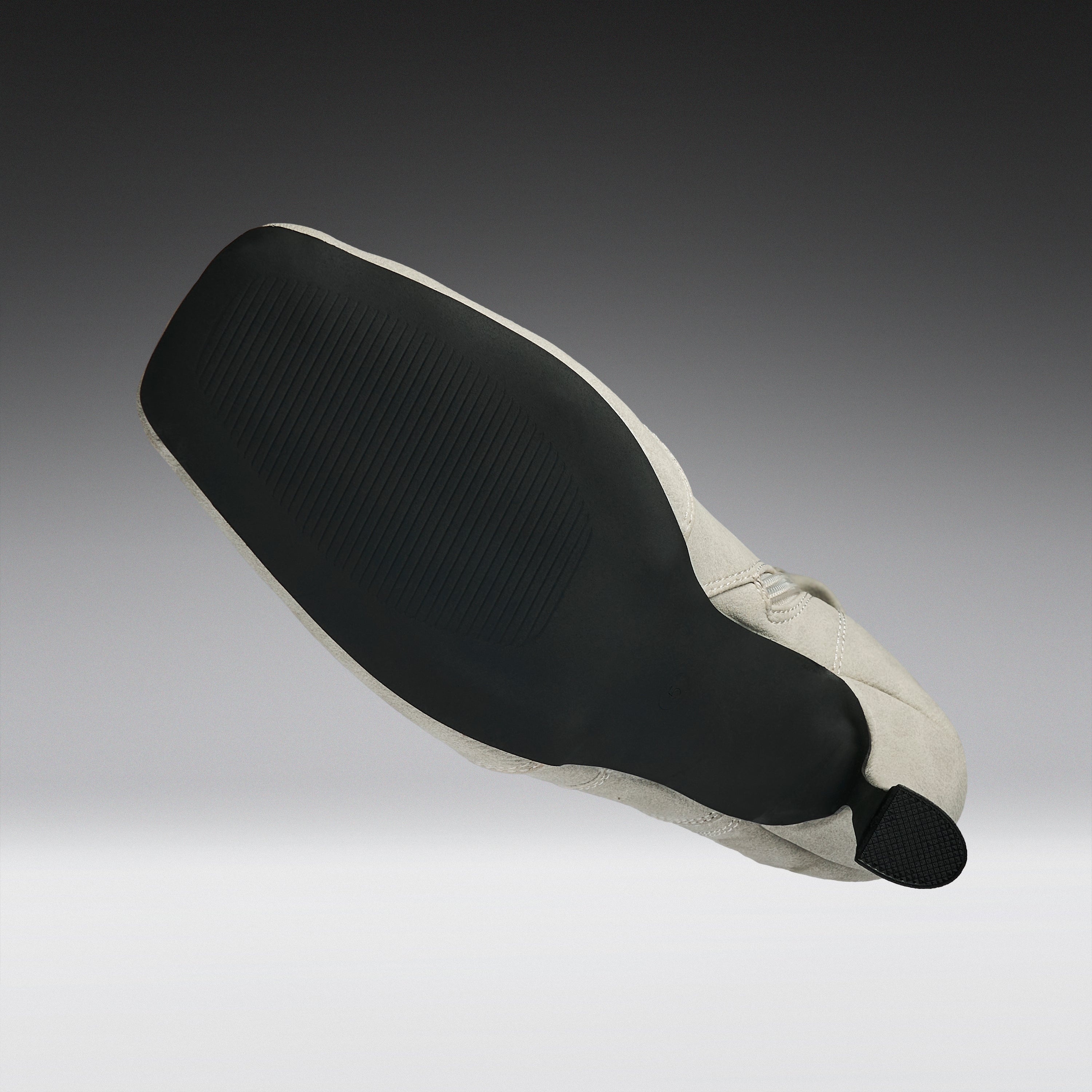 A close-up of a shoe's outsole and heel area, showing a black textured rubber tread and a light beige fabric heel cover with a small black pull tab, set against a dark-to-light gray gradient background.
