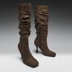 A pair of brown suede slouchy knee-high boots with kitten heels, shown on a neutral gray gradient studio background.