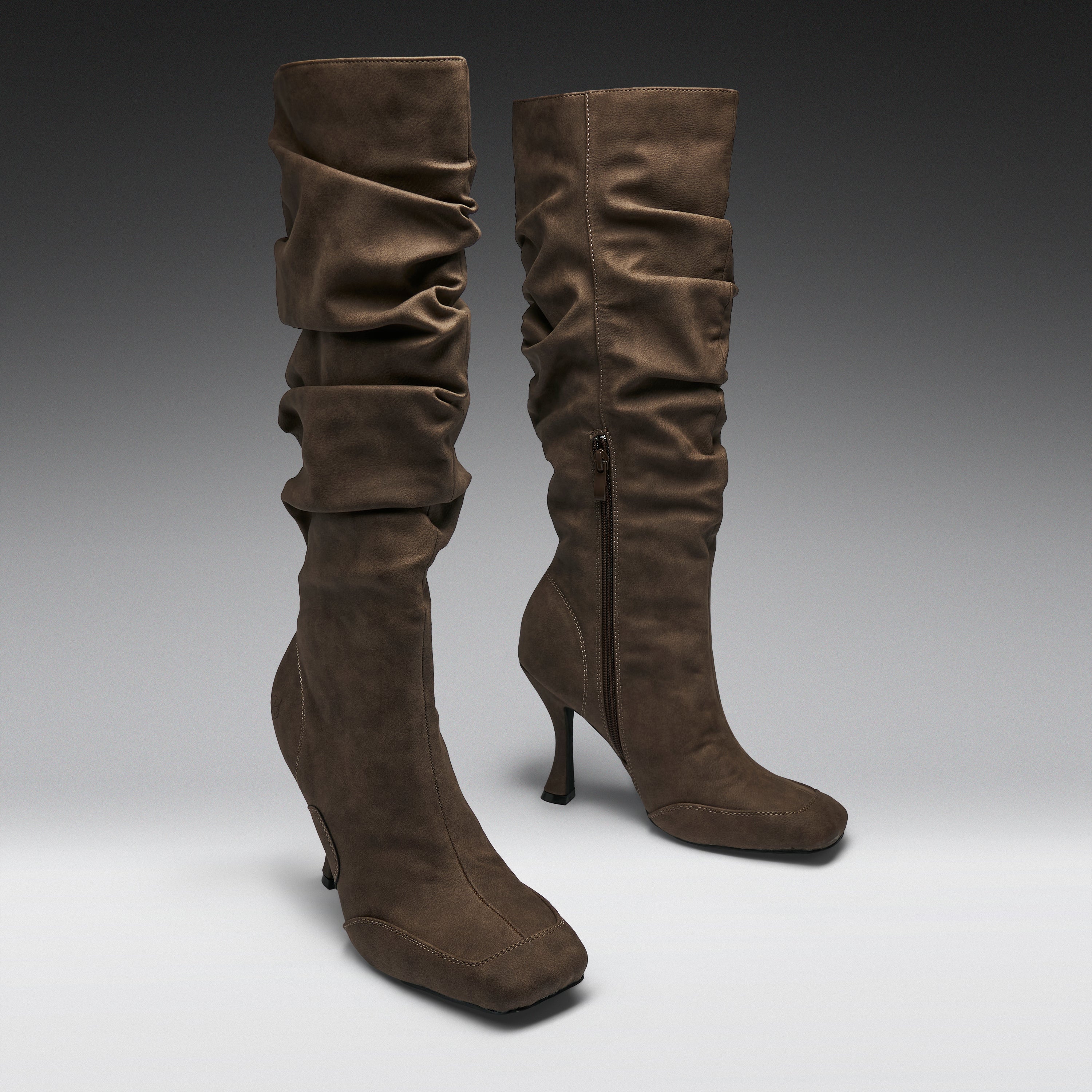 A pair of brown suede slouchy knee-high boots with kitten heels, shown on a neutral gray gradient studio background.