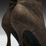 Close-up of a brown suede high-heeled boot showing the side heel, stitching, and a small embossed logo on a dark gray gradient background.