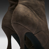 Close-up of a brown suede high-heeled boot showing the side heel, stitching, and a small embossed logo on a dark gray gradient background.